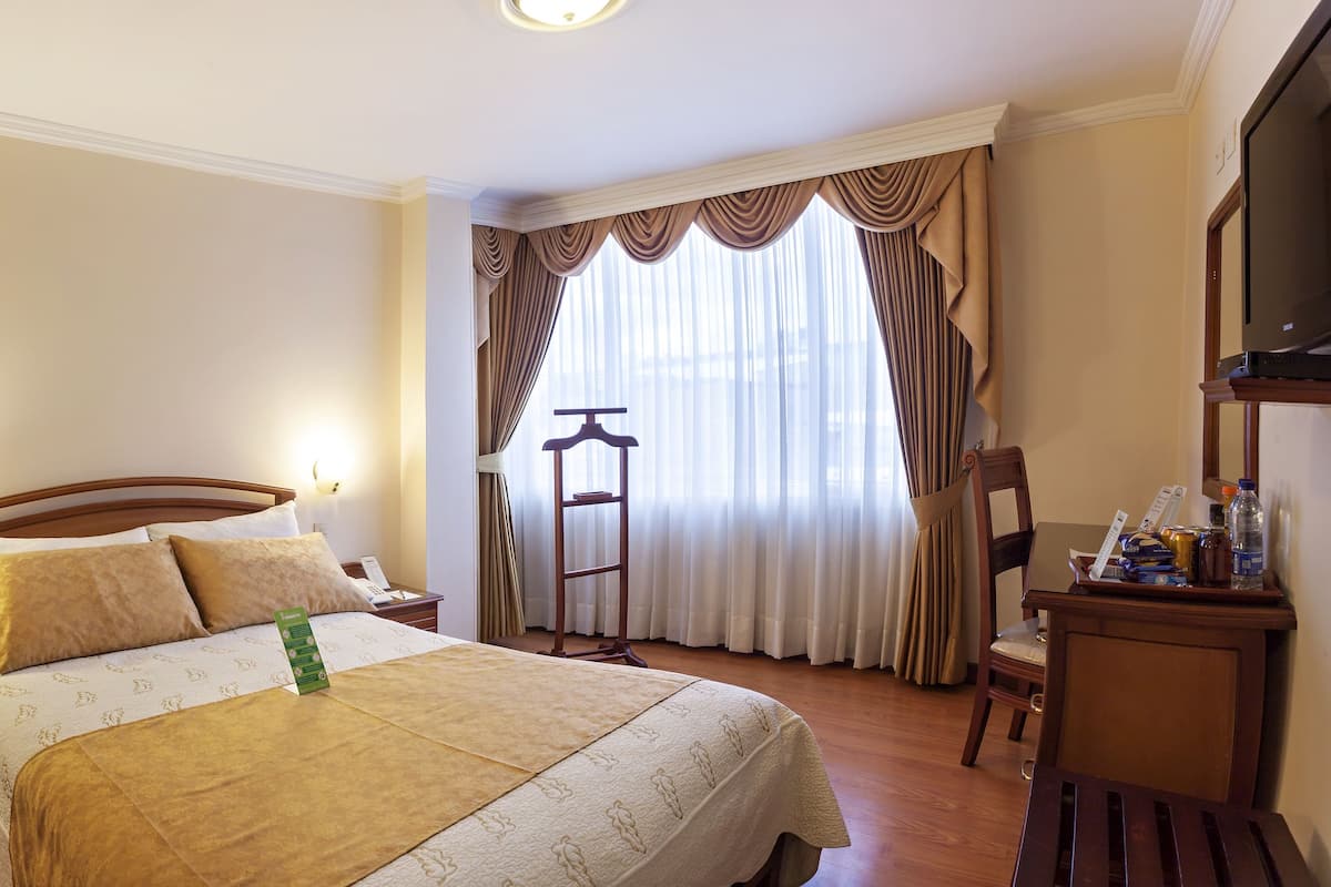 A hotel room with a bed, bedside table, nightstand, and a window with curtains.