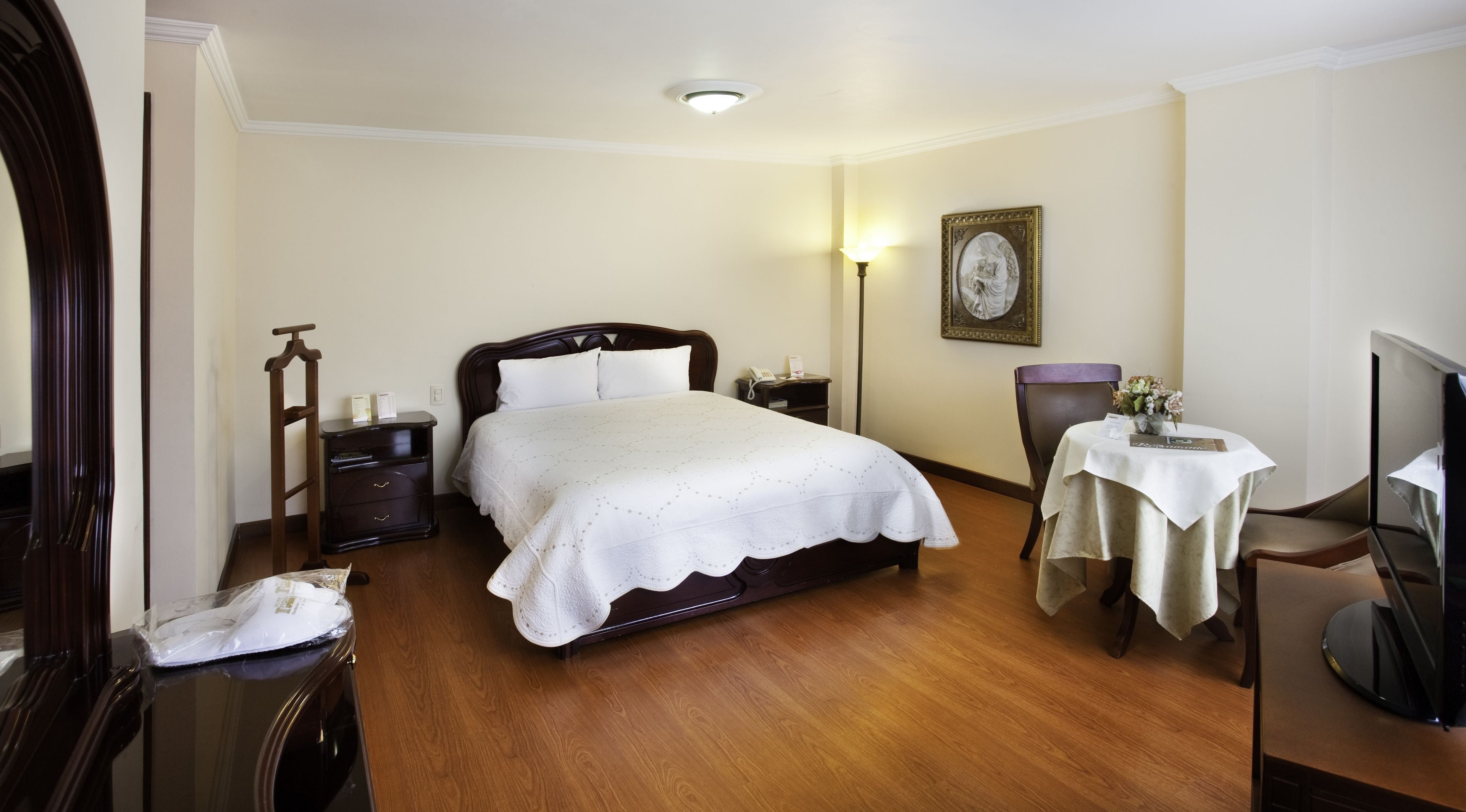 A hotel room with a large bed, bedside table, lamp, chair, table, and a television.