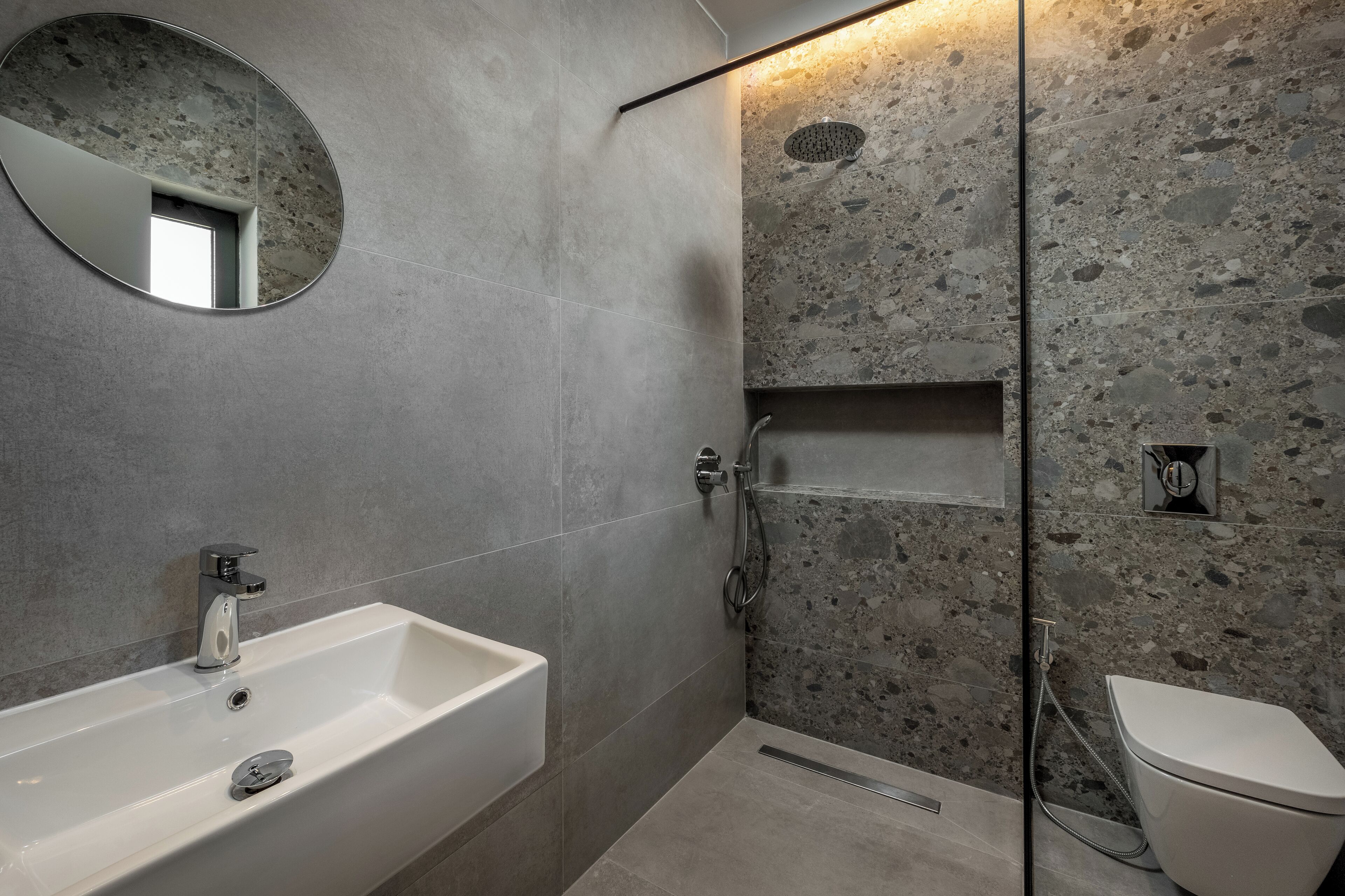 Un baño moderno con un lavabo blanco, un espejo redondo, ducha con pared de piedra y un inodoro.