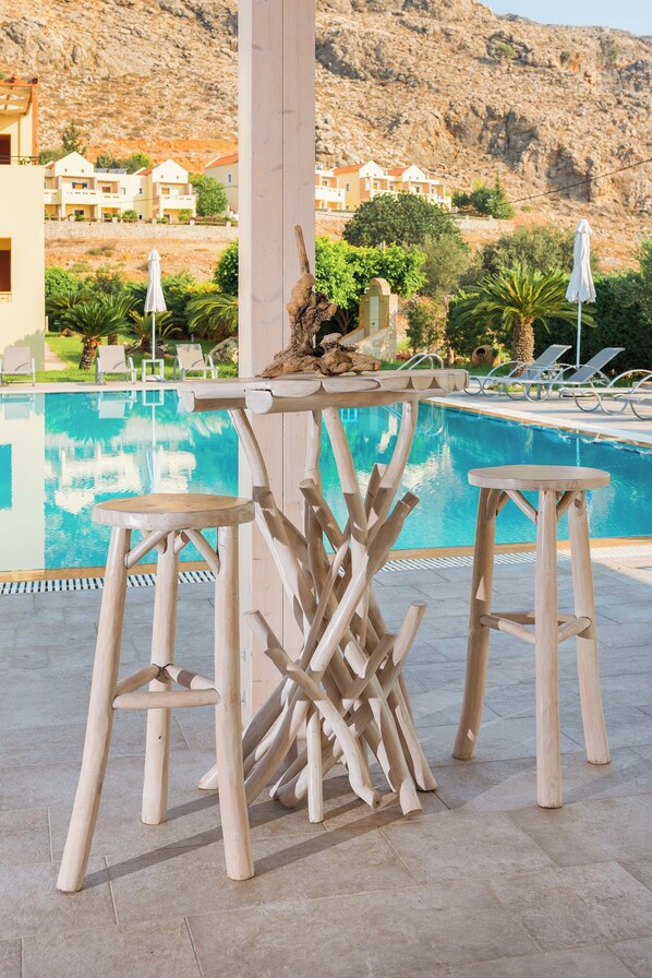A poolside area with wooden stools and a bar, overlooking a pool and a mountainous backdrop. - Celestial Lindos suites (Rhodes)