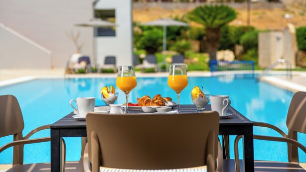 Una mesa con alimentos para el desayuno y dos vasos de jugo de naranja junto a una piscina, con vistas a una zona residencial.