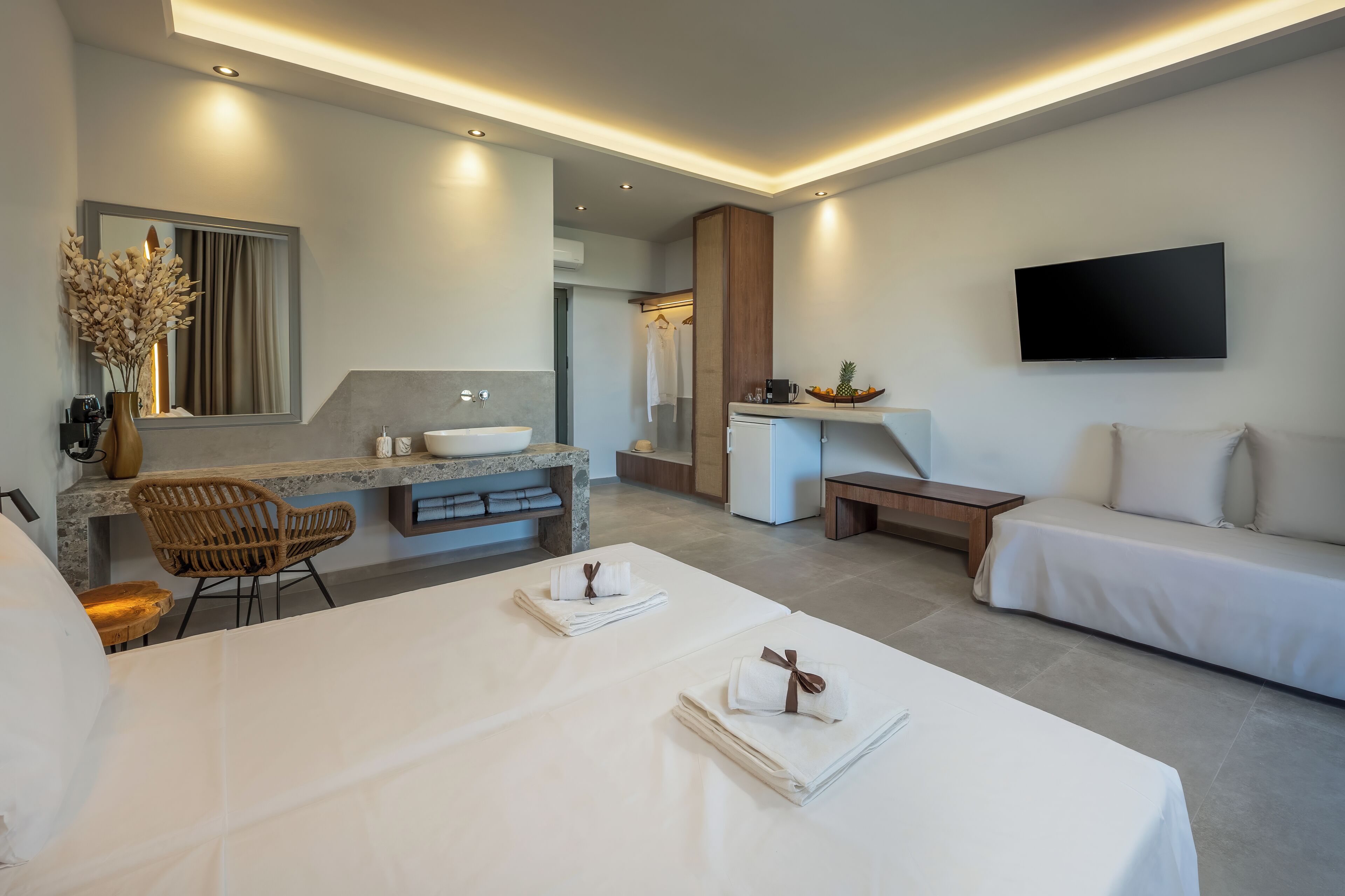 Ein modernes Hotelzimmer mit einem großen Bett, einer Sitzecke inklusive Sofa und kleinem Tisch, einem Badezimmer mit Waschbecken und Spiegel sowie einem an der Wand montierten Flachbildfernseher.