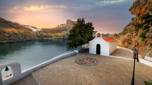 Escena costera con un pequeño edificio blanco, un área pavimentada con un mosaico circular y un litoral rocoso.