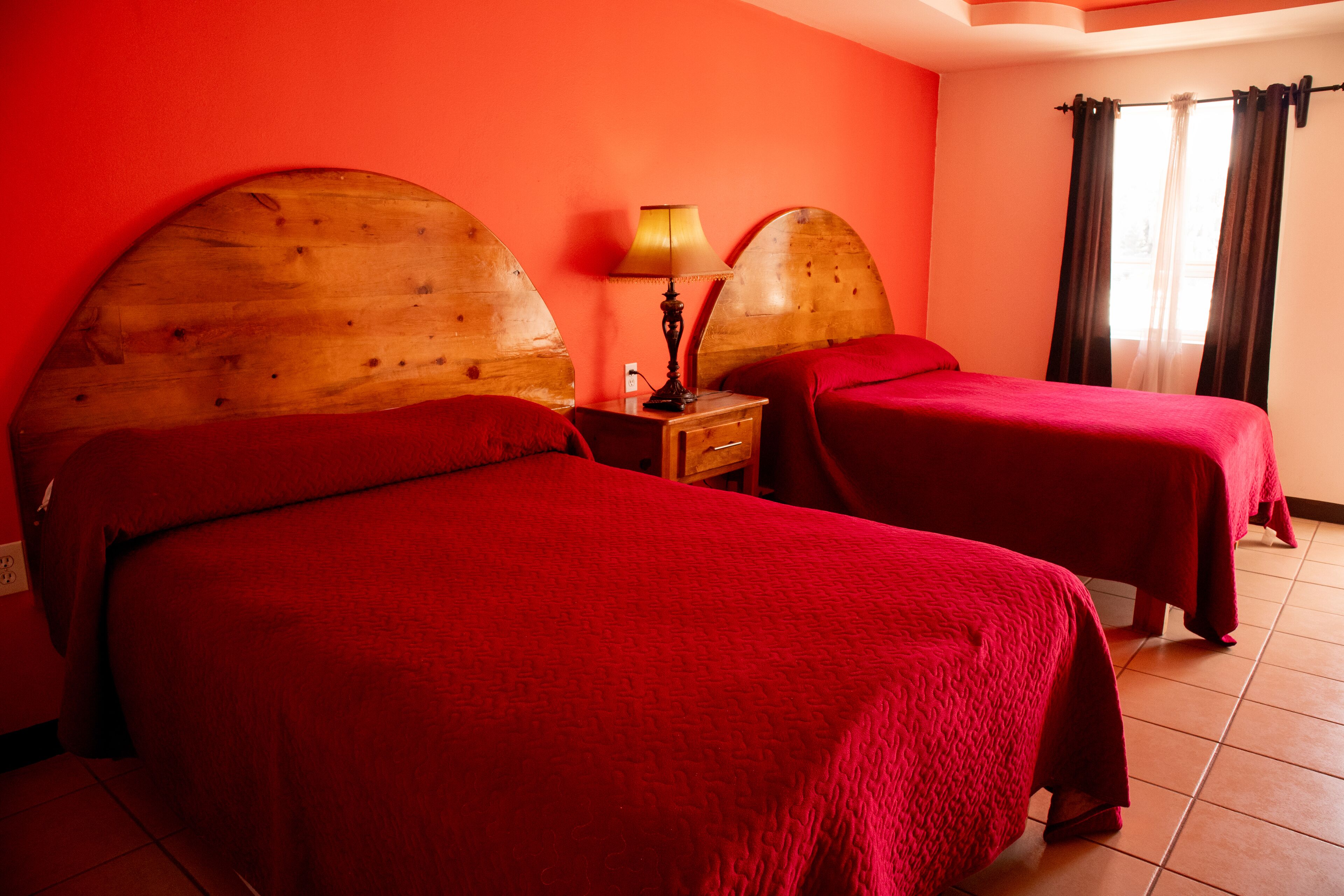 A room with two beds, a nightstand with a lamp, and a window with curtains.