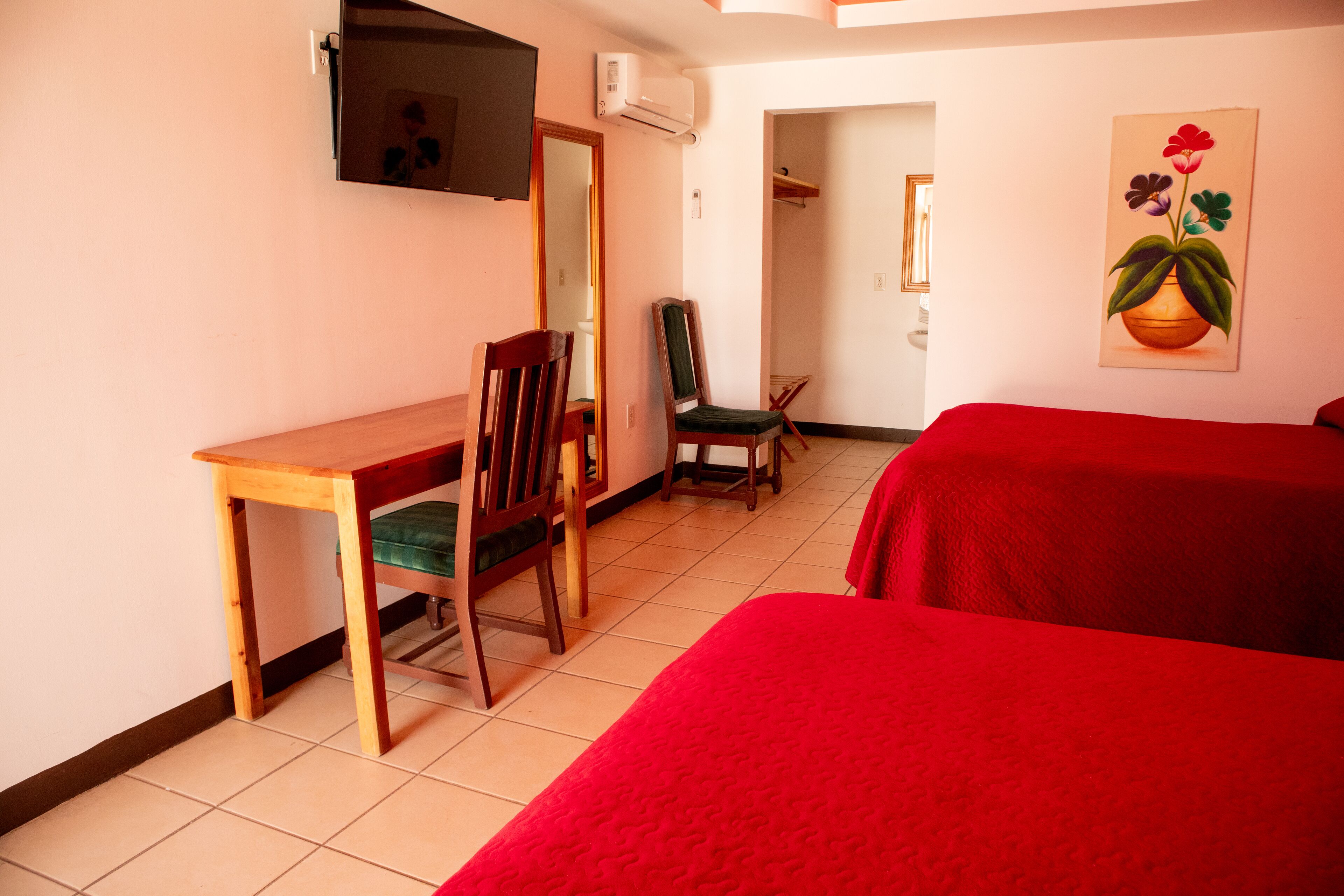 A room with a wooden desk, two chairs, a bed with red bedding, a TV mounted on the wall, and a painting of flowers.