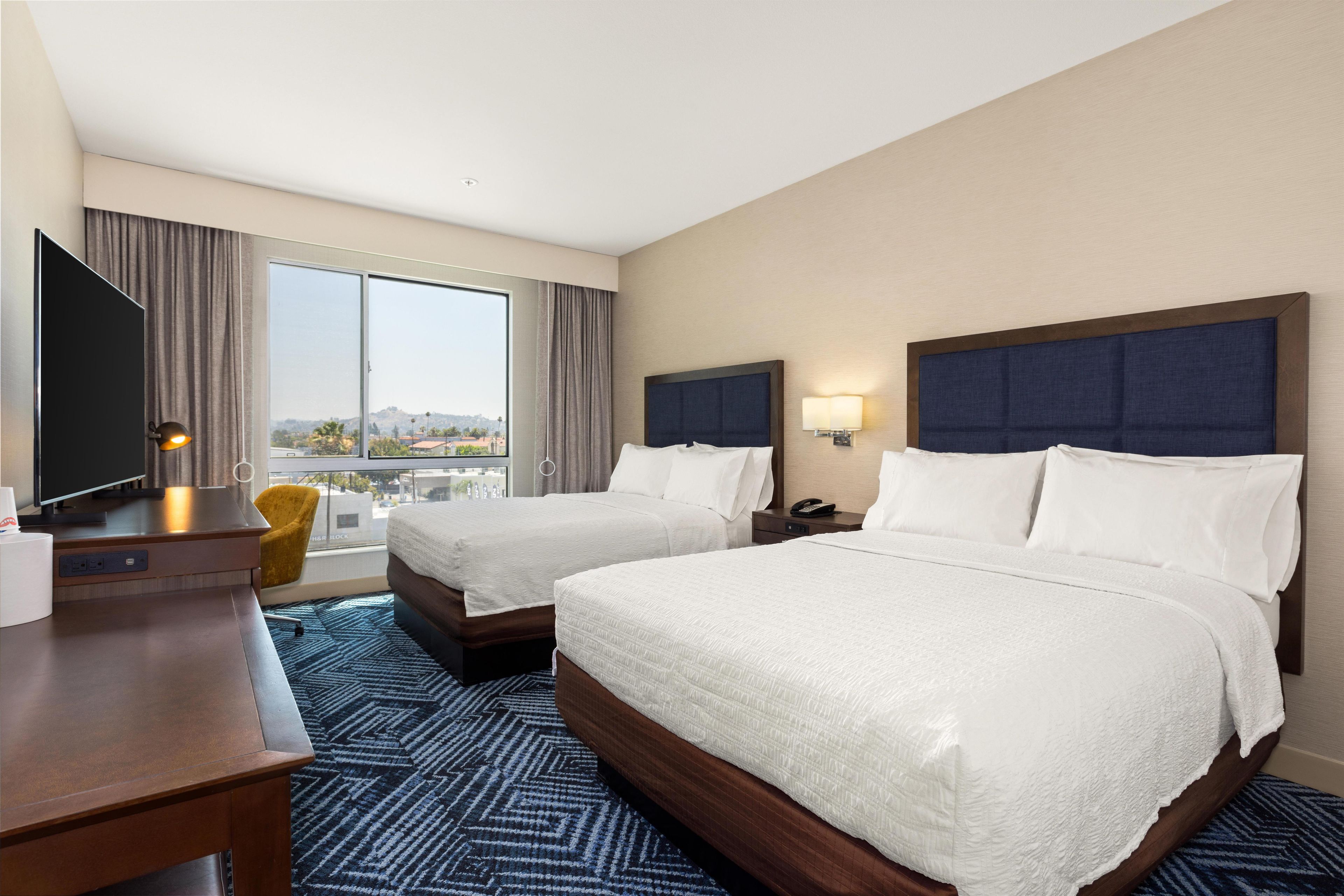 A hotel room with two beds, a TV, a desk, a chair, and a large window with a city view.