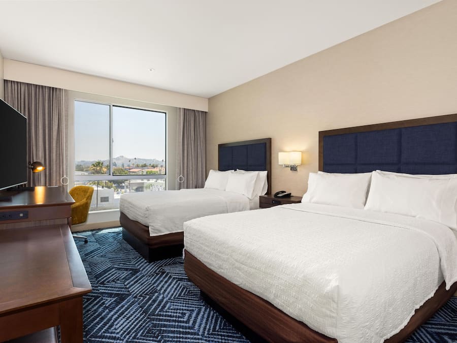 A hotel room with two beds, a TV, a desk, a chair, and a large window with a city view.