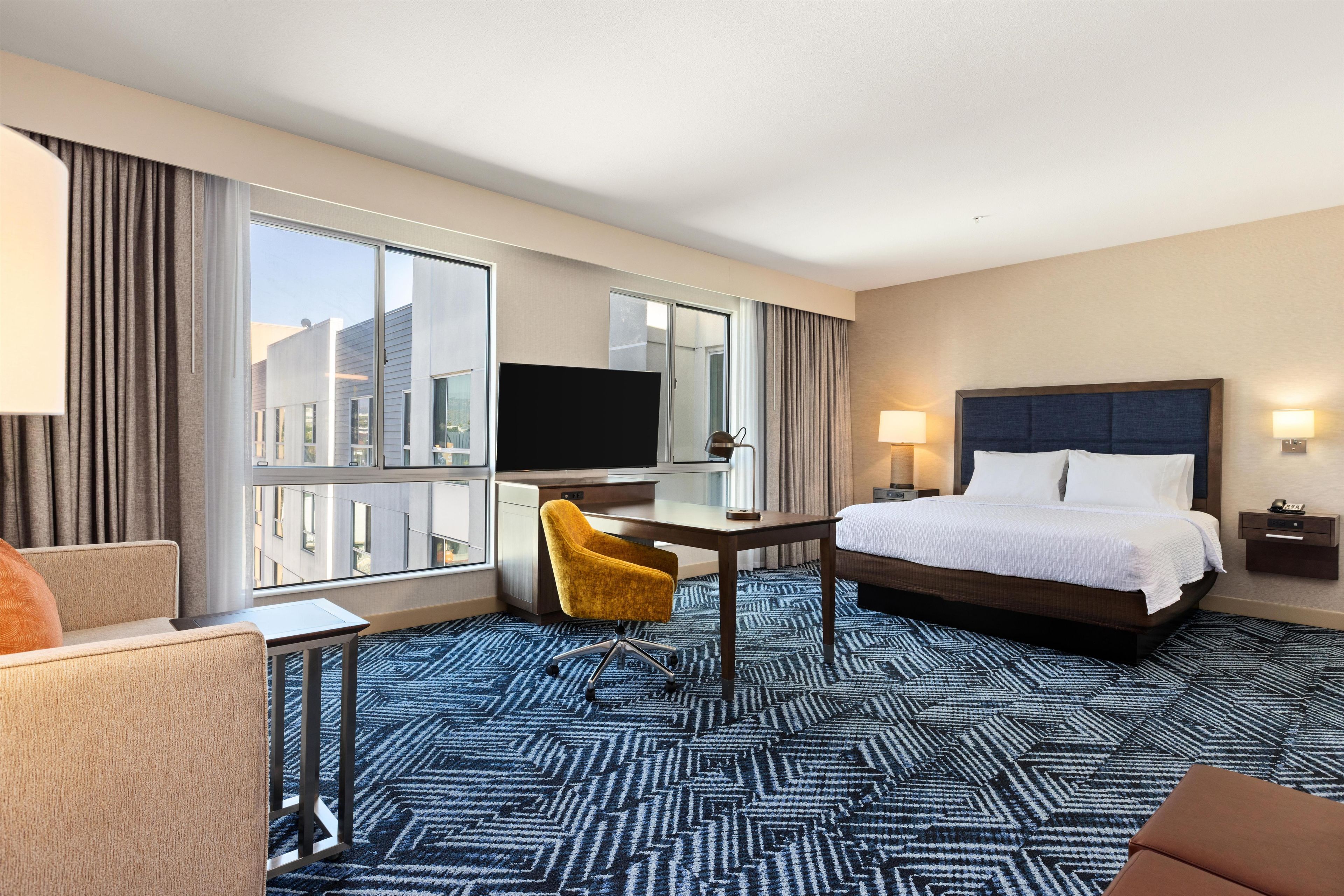 A hotel room with a bed, a desk with a TV, a chair, a large window, and a view of buildings.