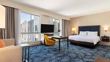 A hotel room with a bed, a desk with a TV, a chair, a large window, and a view of buildings.
