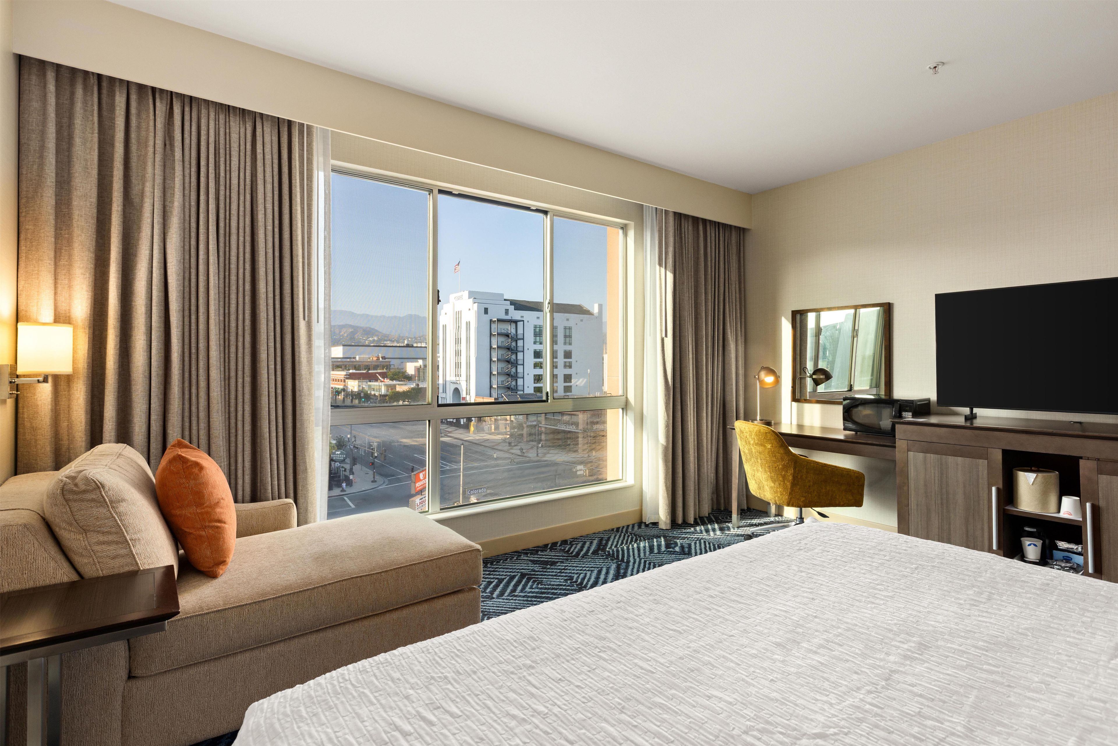 A hotel room with a large bed, a desk, a chair, and a view of a cityscape through the window.