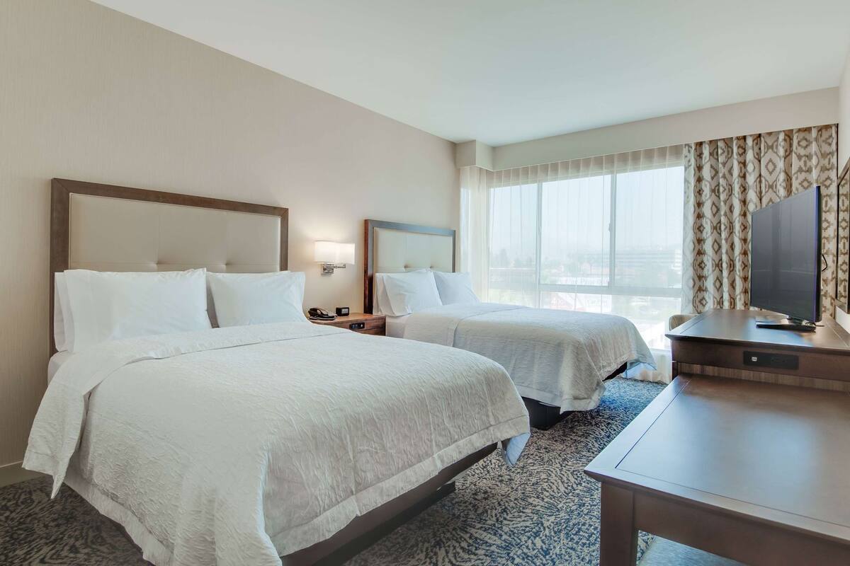 A hotel room with two beds, a TV, a desk, and a large window with curtains.