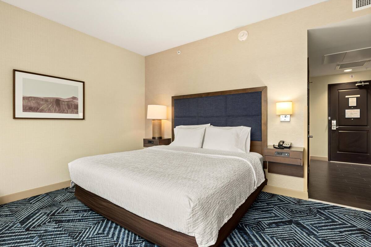 A hotel room with a bed, two bedside tables, a nightstand, a framed picture on the wall, and a door to the right.