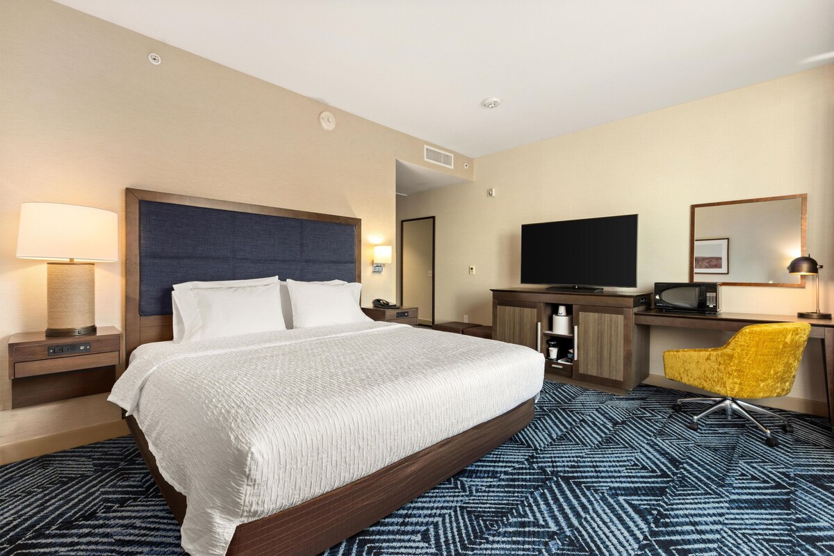 A hotel room with a large bed, a desk with a flat-screen TV, a yellow chair, and a patterned carpet.