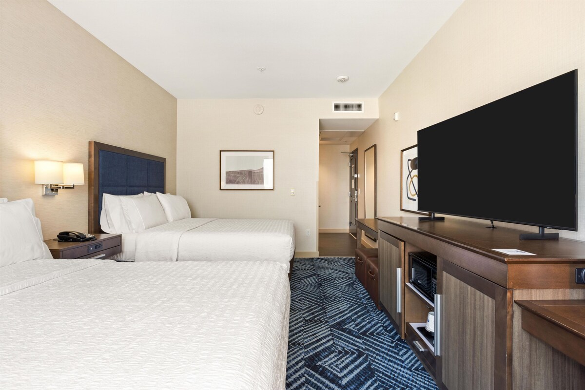 A hotel room with two beds, a flat-screen TV, a desk with a lamp, and a framed picture on the wall.