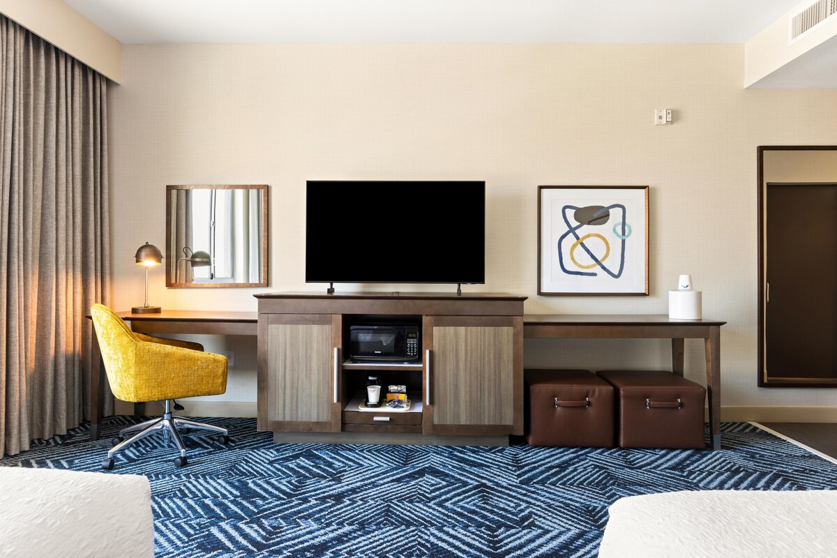 A hotel room with a bed, a desk with a chair, a television, a coffee maker, and a framed abstract artwork.