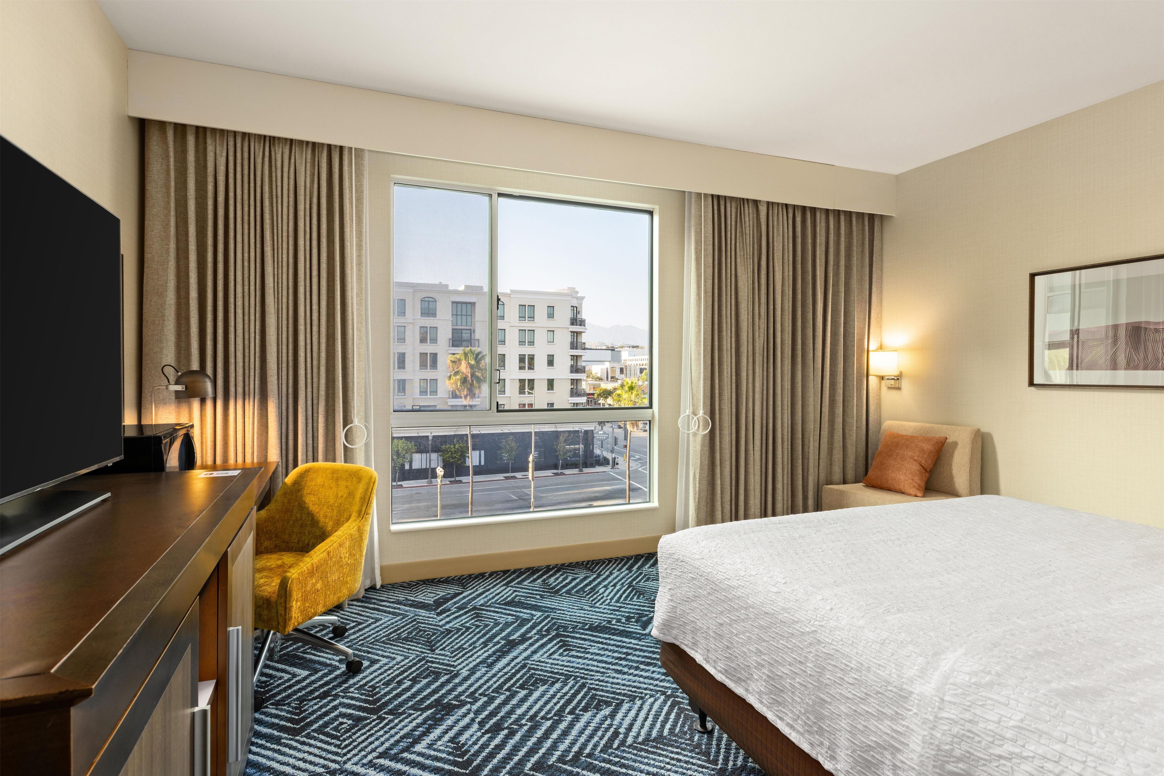 Hotelkamer met een bed, bureau, stoel, televisie en een groot raam met gordijnen.