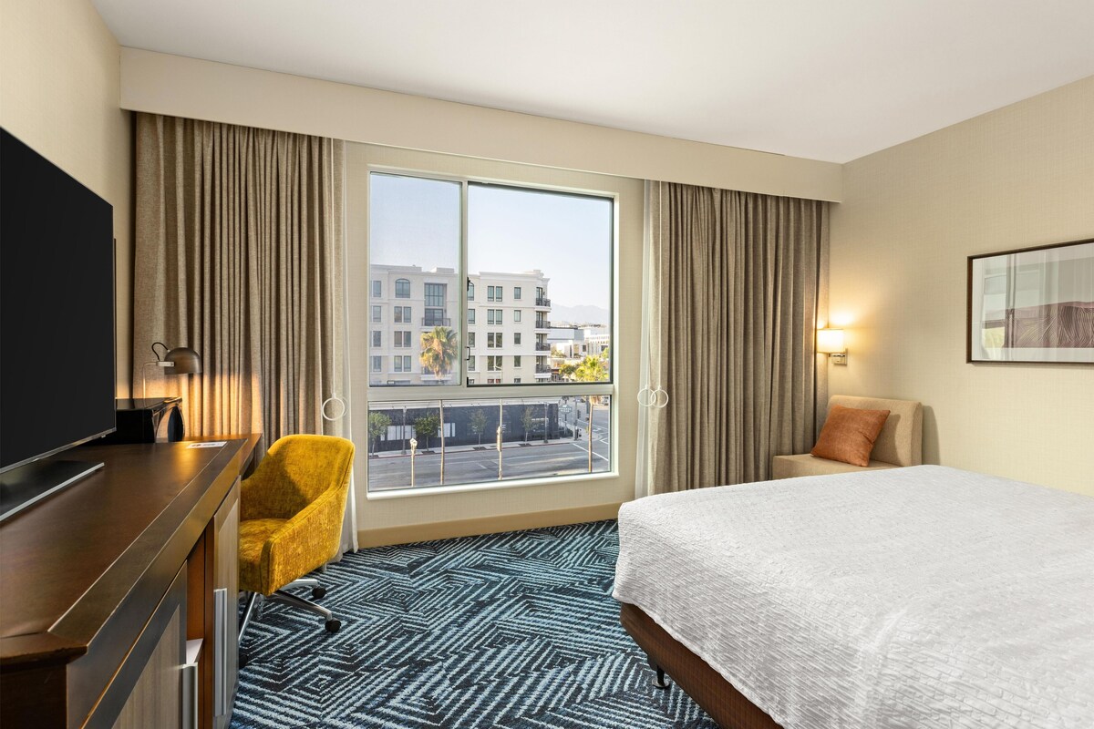 A hotel room with a bed, a desk, a chair, a TV, and a large window with curtains.