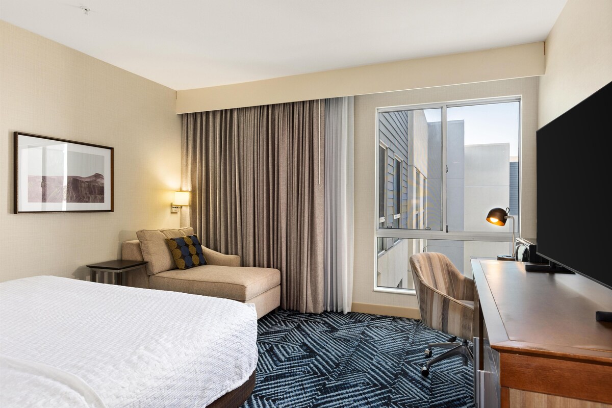 A hotel room with a bed, a sofa, a desk with a chair, a television, and a window with curtains.