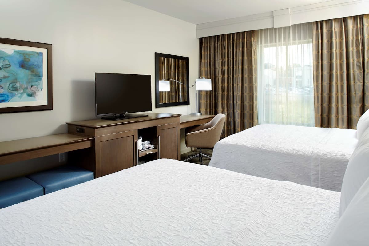 A hotel room with two beds, a desk with a TV, a chair, a mirror, and a window with curtains.