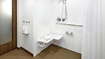 Un baño moderno con pared de azulejos blancos, puerta de madera, lavamanos blanco con toallero, ducha con cabezal de ducha manual y retrete con barra de apoyo.