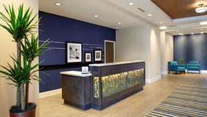 A reception area with a dark blue wall, a reception desk, framed pictures, a potted plant, and teal chairs. - Hampton Inn & Suites Pittsburgh Airport South–Settlers Ridge (Pittsburgh)