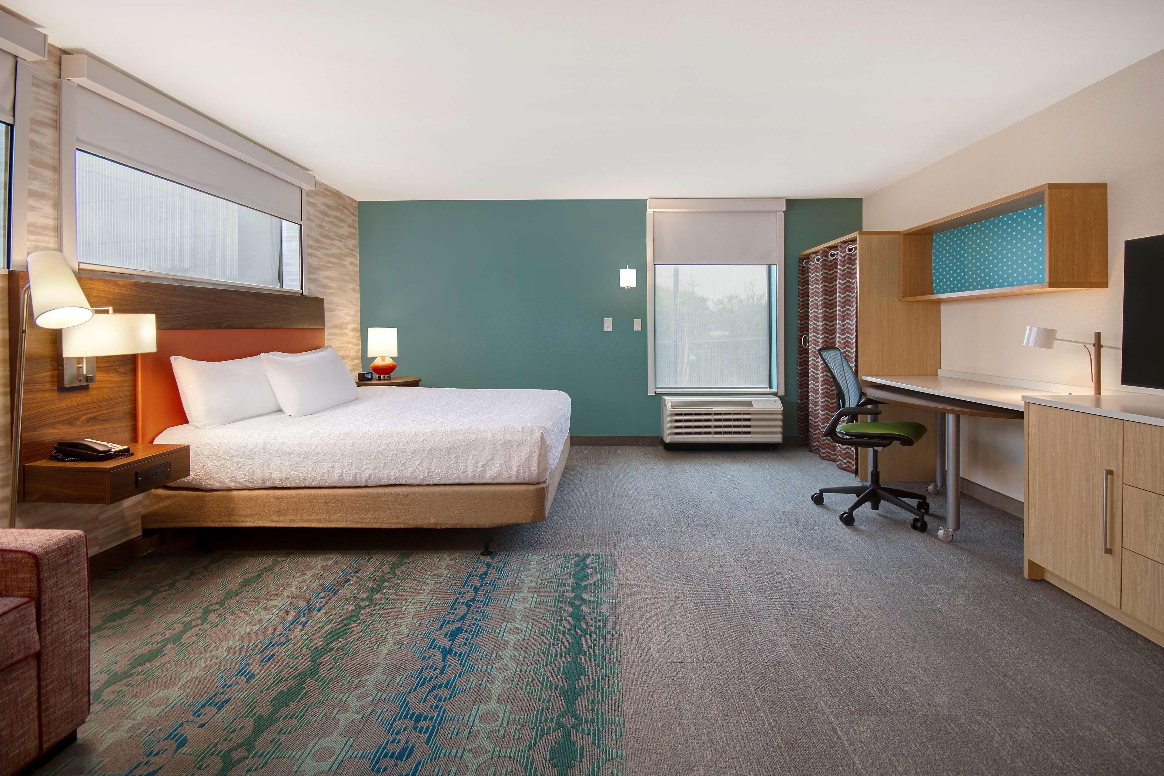 A hotel room with a bed, desk, and chair, a window, and an air conditioning unit.