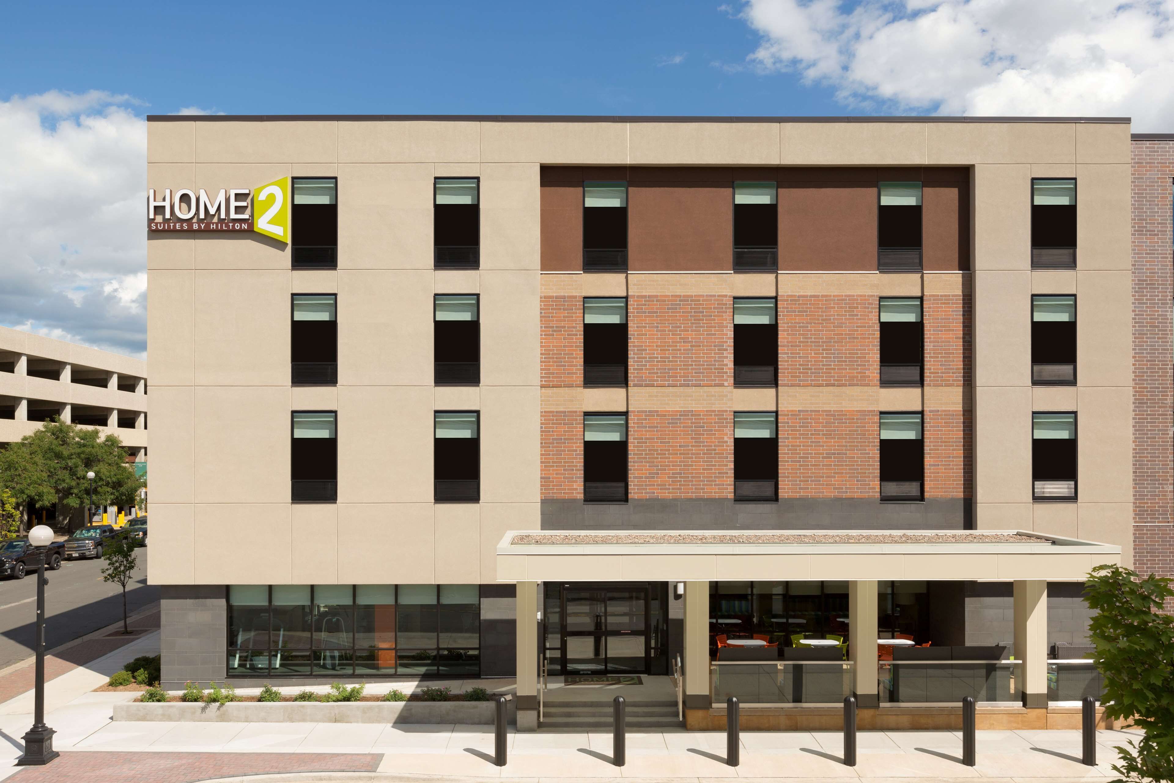 Un moderno edificio de hotel con varios pisos, un letrero llamativo que dice "HOME 2 Suites by Hilton" y un área de entrada cubierta.