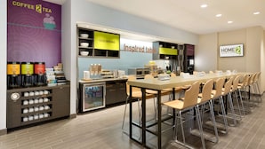 Un hall d’hôtel moderne avec un coin café, une table au design original et un panneau « Home2 ».