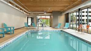 Une piscine intérieure à l’eau claire, entourée de chaises bleues et de chaises orange, avec de grandes fenêtres donnant sur l’extérieur.