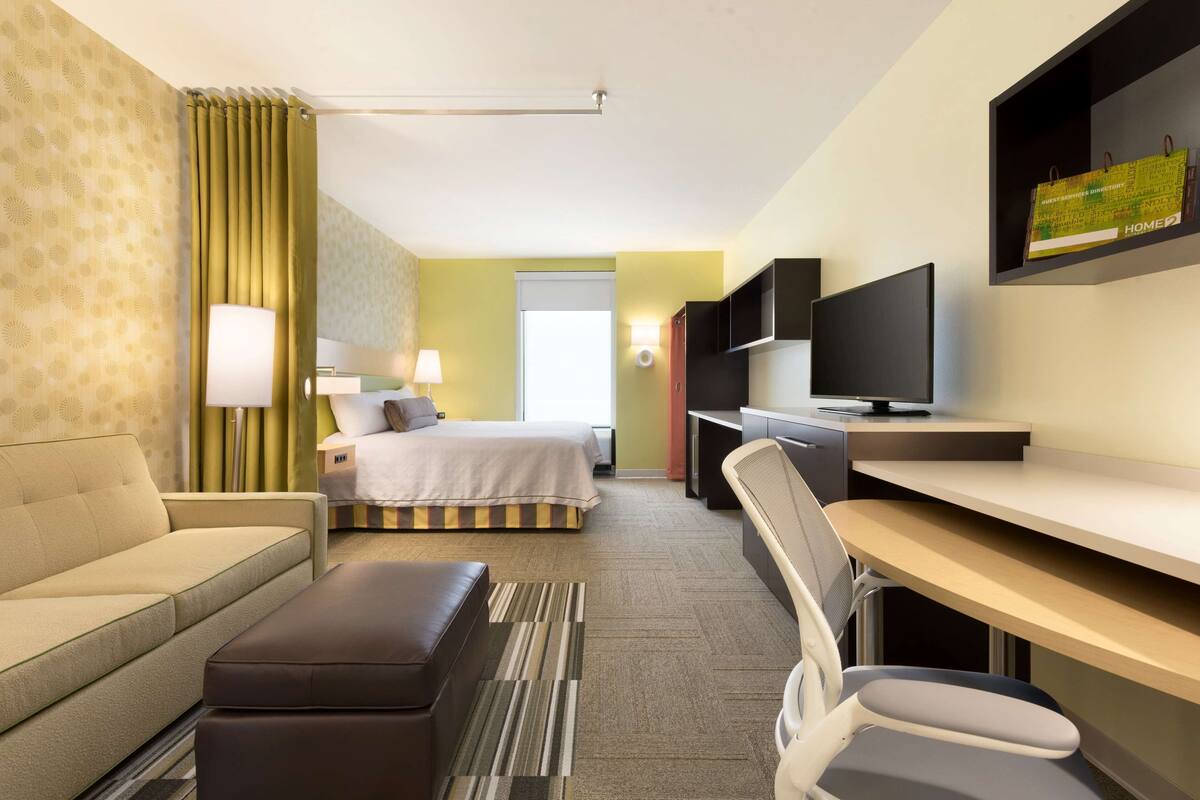 A hotel room with a bed, a sofa, a desk, a chair, a TV, and a lamp.