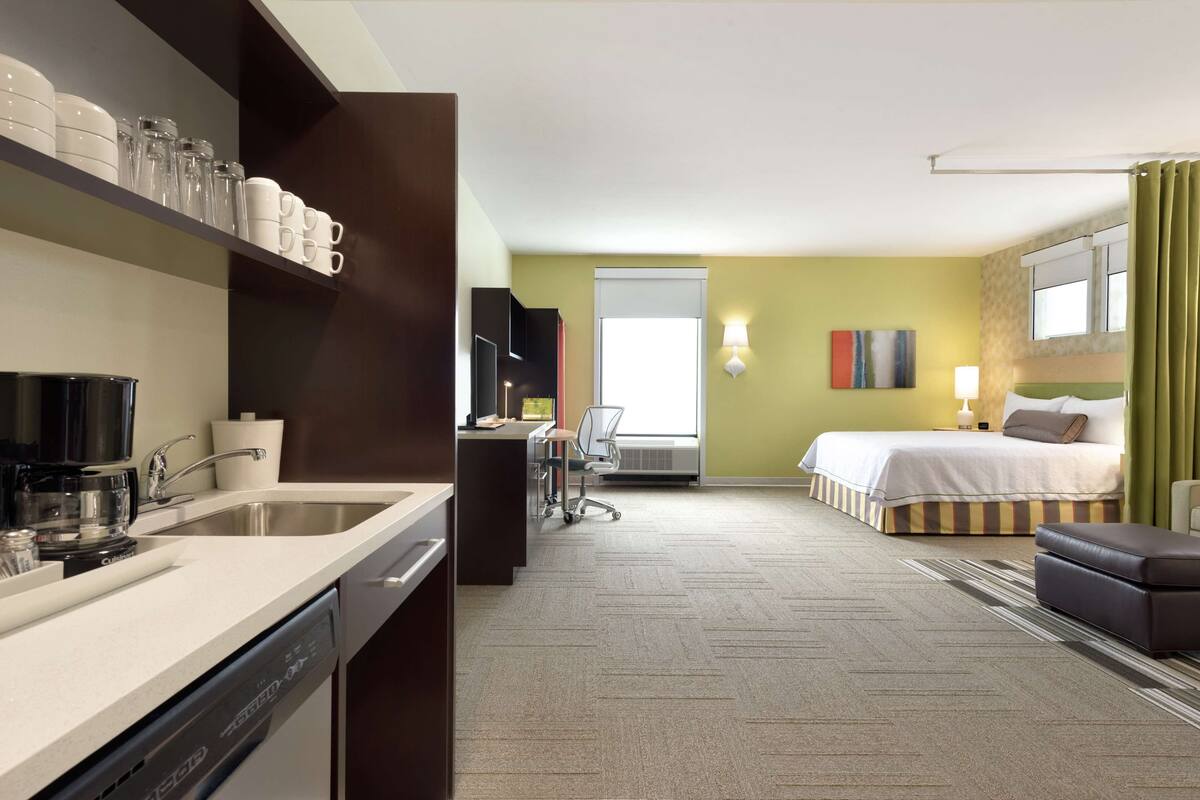 A hotel room with a kitchen area, a bed, a desk, and a chair.