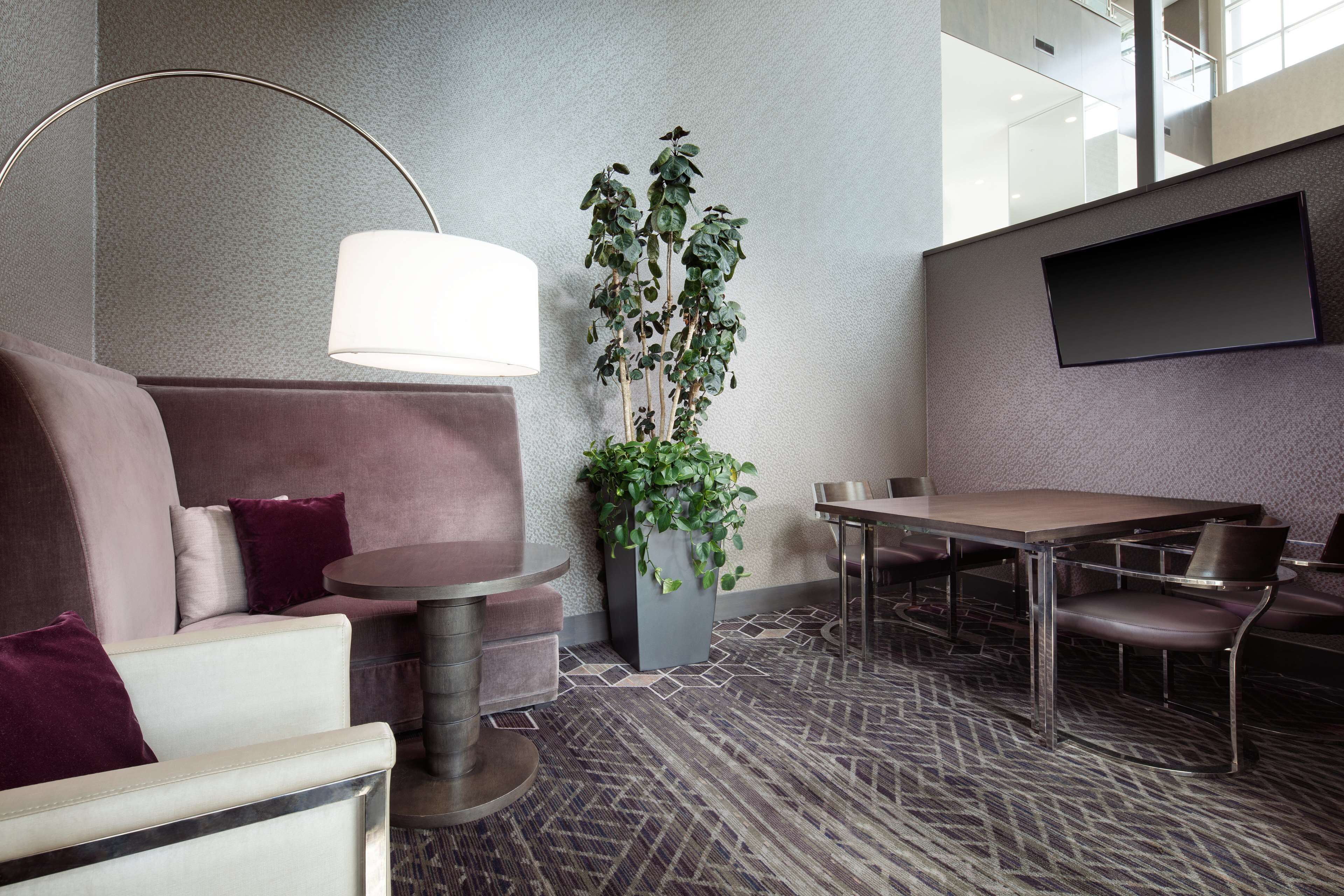 Una habitación de hotel moderna con sofá, una mesa redonda, una mesa de comedor, una planta en maceta y un televisor de pantalla plana montado en la pared.
