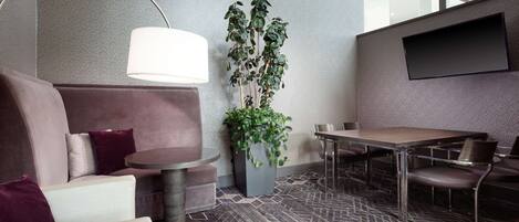 Una habitación de hotel moderna con sofá, una mesa redonda, una mesa de comedor, una planta en maceta y un televisor de pantalla plana montado en la pared.