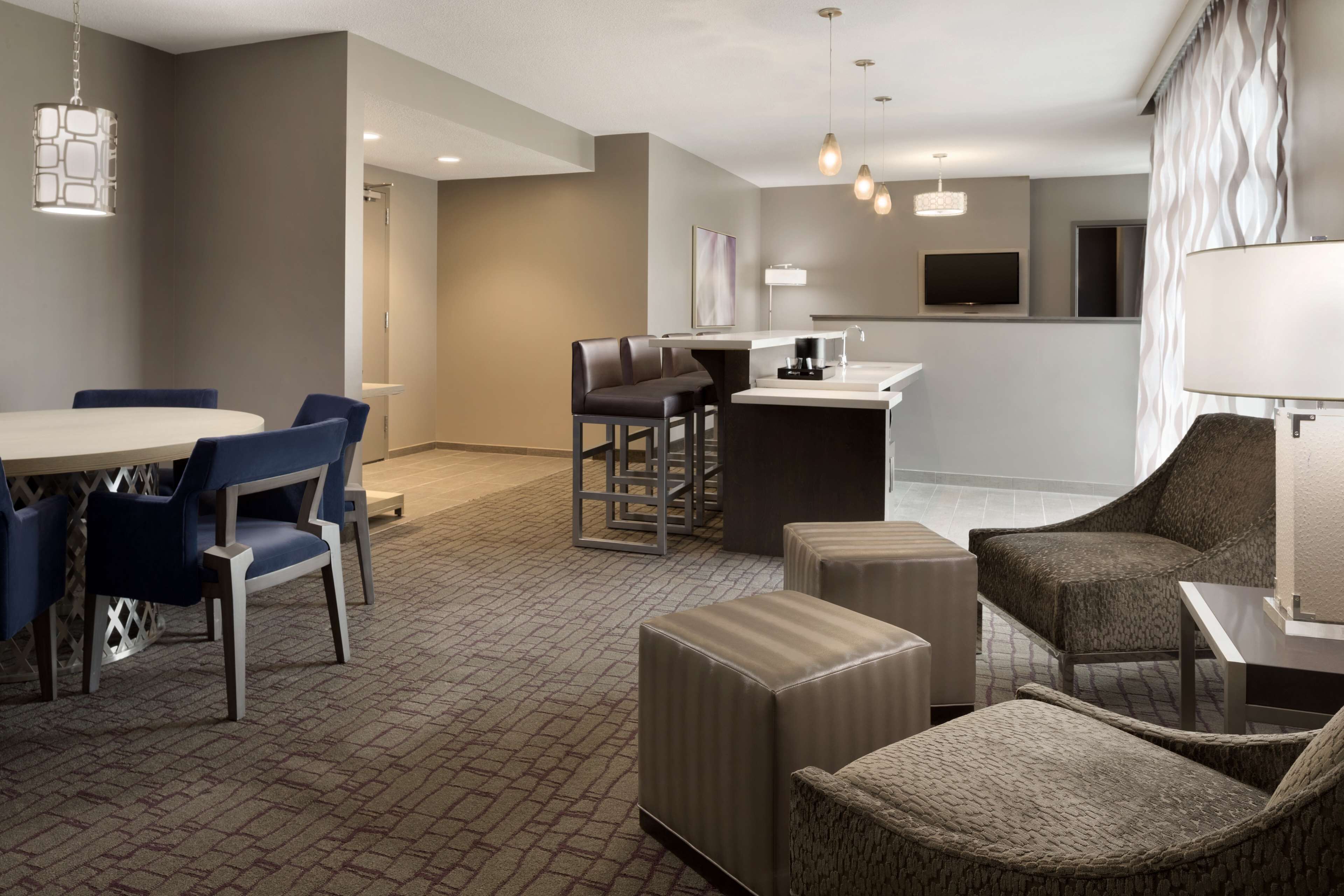 A modern hotel room with a dining area, a kitchen counter, and a seating area with ottomans and chairs.