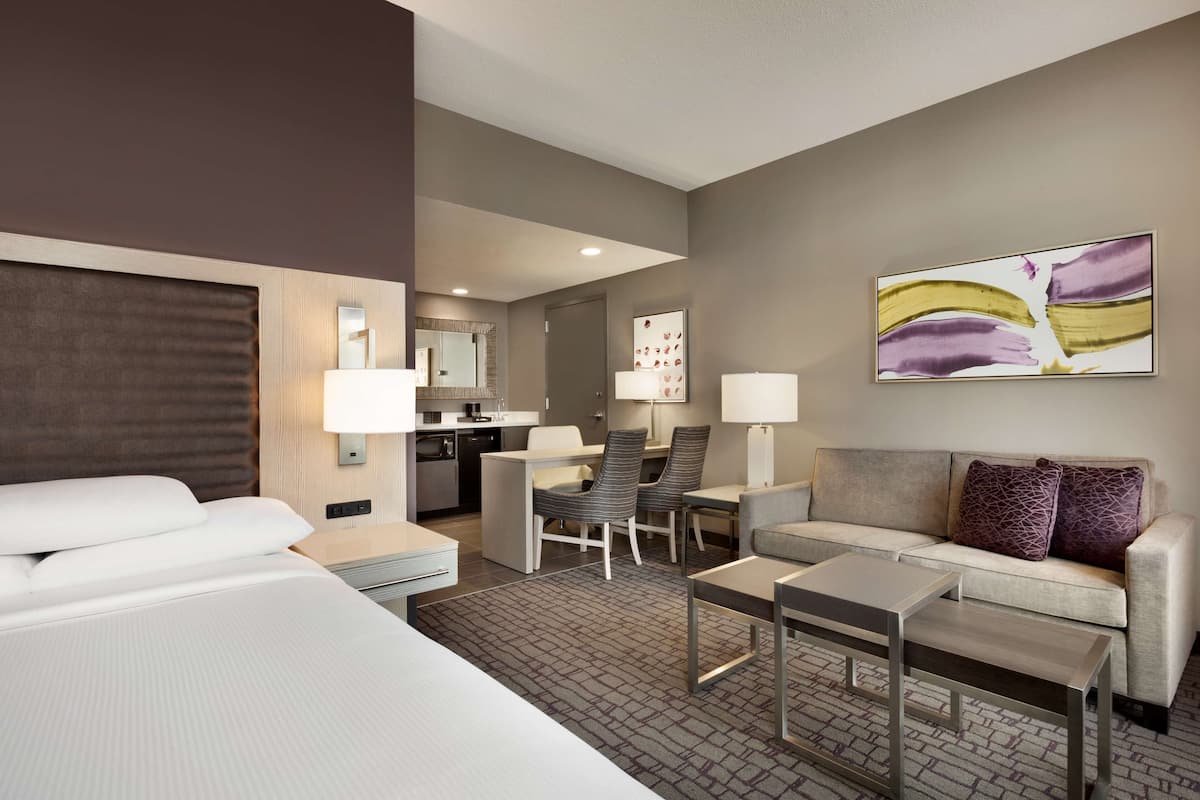 Een hotelkamer met een bed, een slaapbank, een klein tafeltje, een bureau en een stoel. Er is een spiegel, een lamp en een schilderij aan de muur.