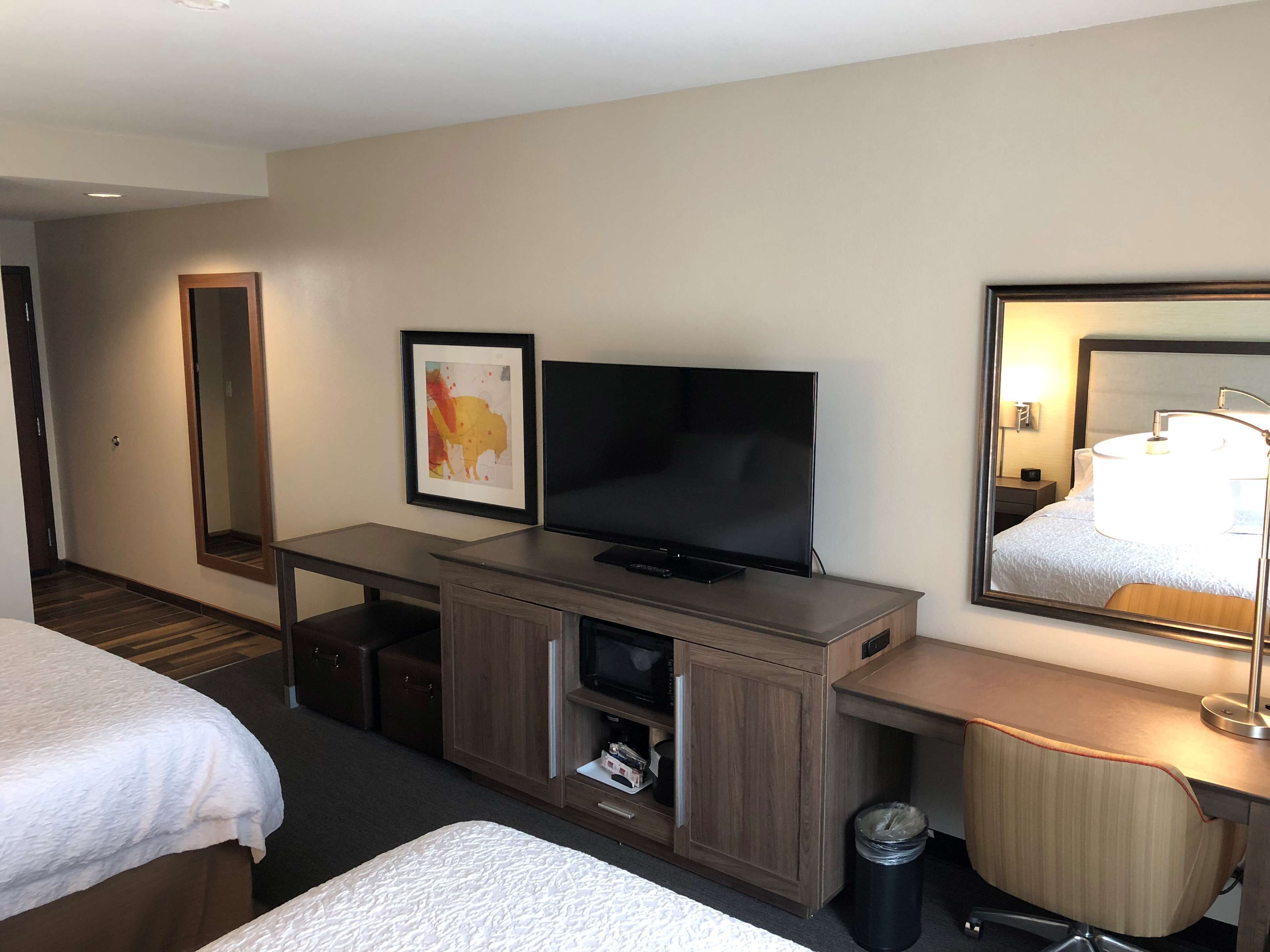 Habitación de hotel con cama, televisión, escritorio, espejo y basurero.