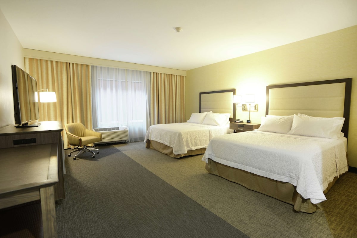 A hotel room with two beds, a desk, a chair, and a large window with curtains.