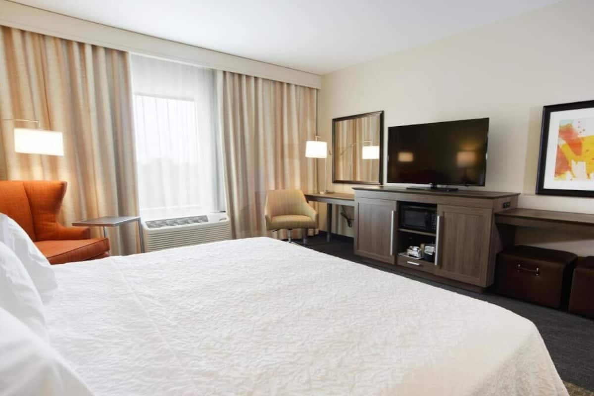 A hotel room with a large bed, a desk with a flat-screen TV, a chair, and a desk lamp.