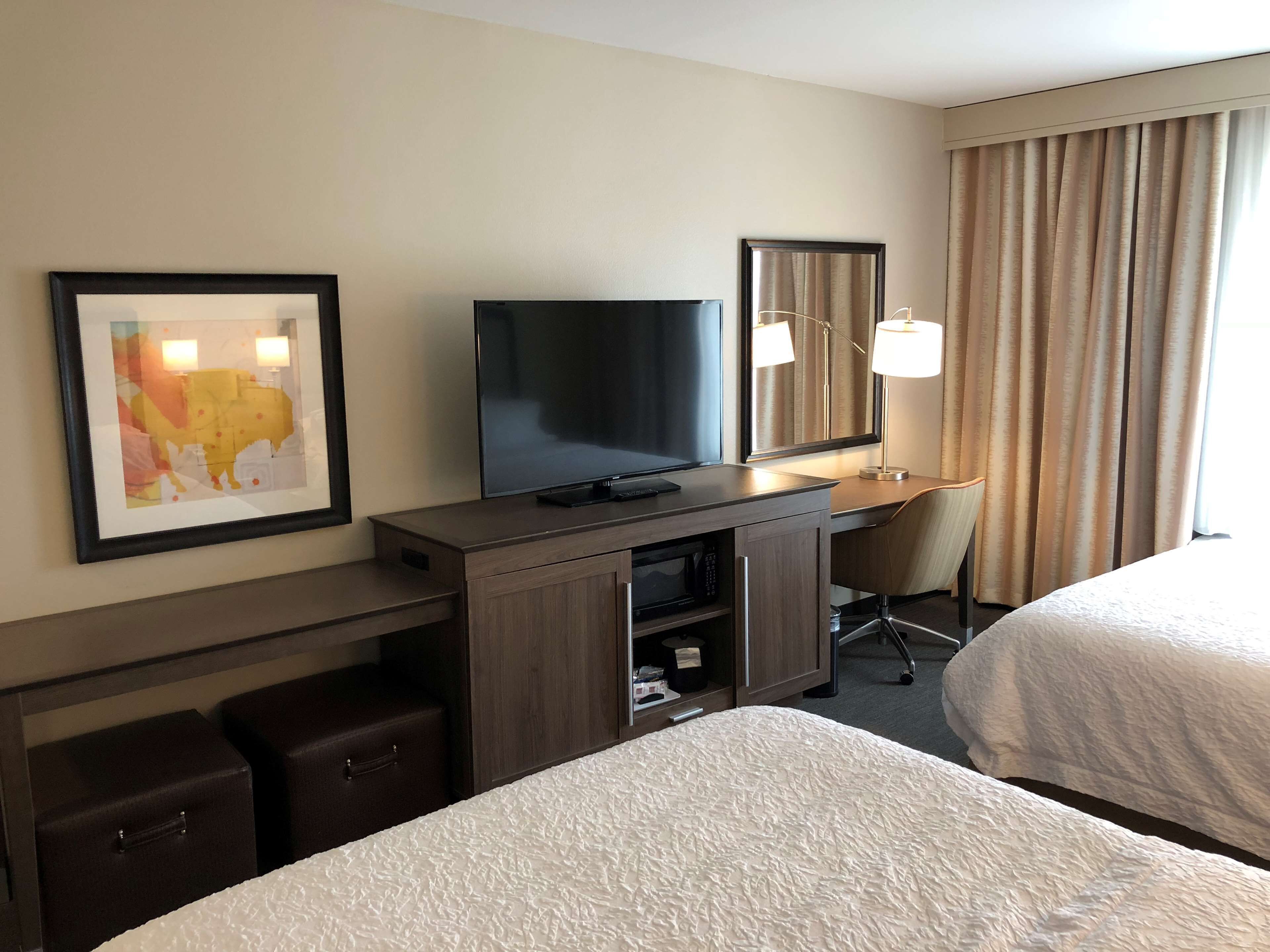 A hotel room with a bed, a desk with a television, a chair, a lamp, and a framed map on the wall.