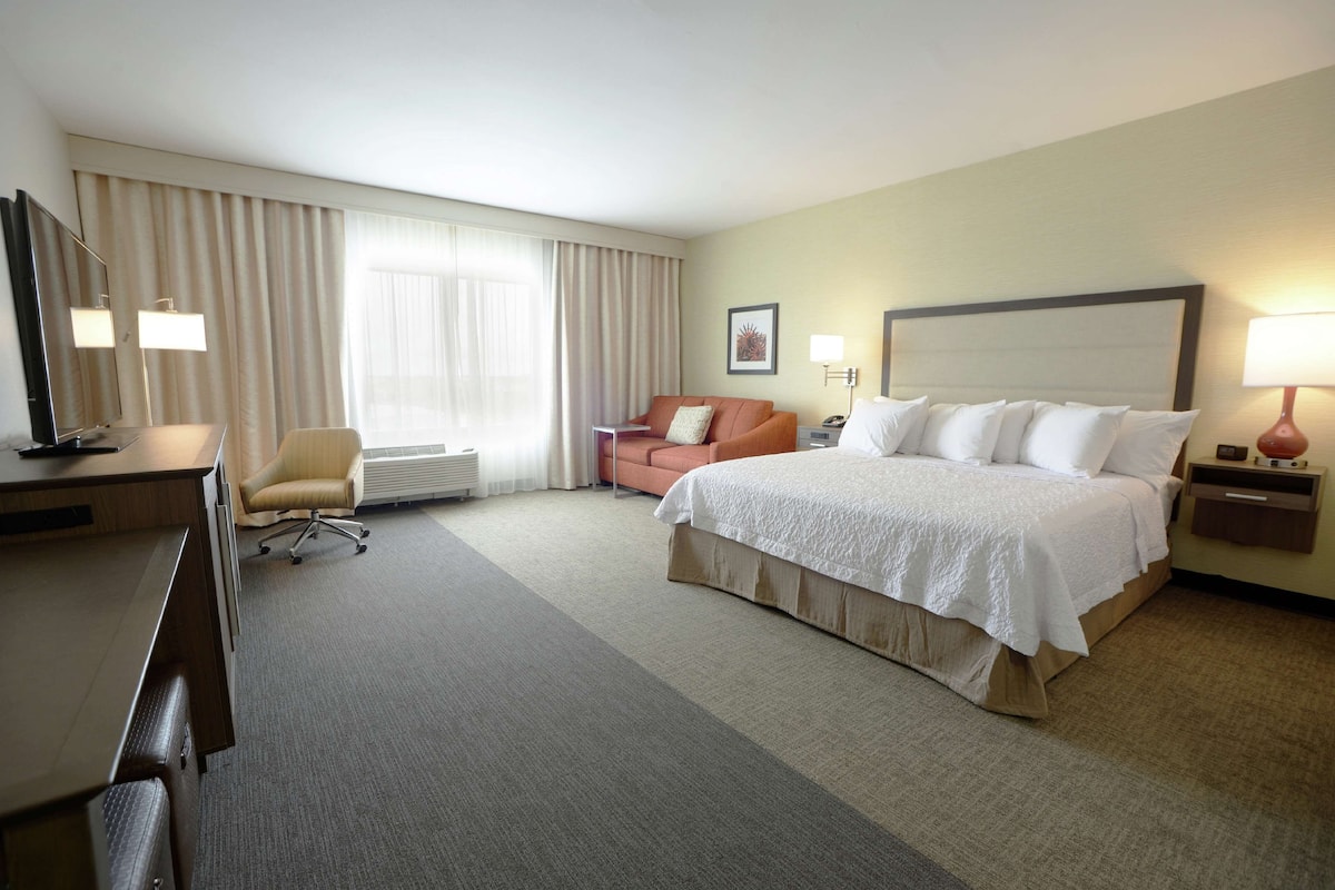 A hotel room with a bed, a desk, a chair, a sofa, and a large window with curtains.