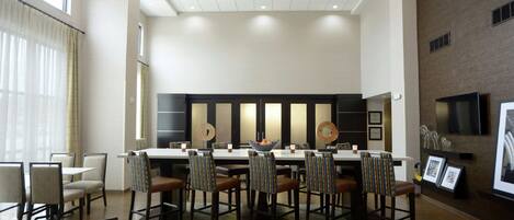 Un espace de salle à manger moderne, avec une longue table, de nombreuses chaises et des luminaires suspendus.