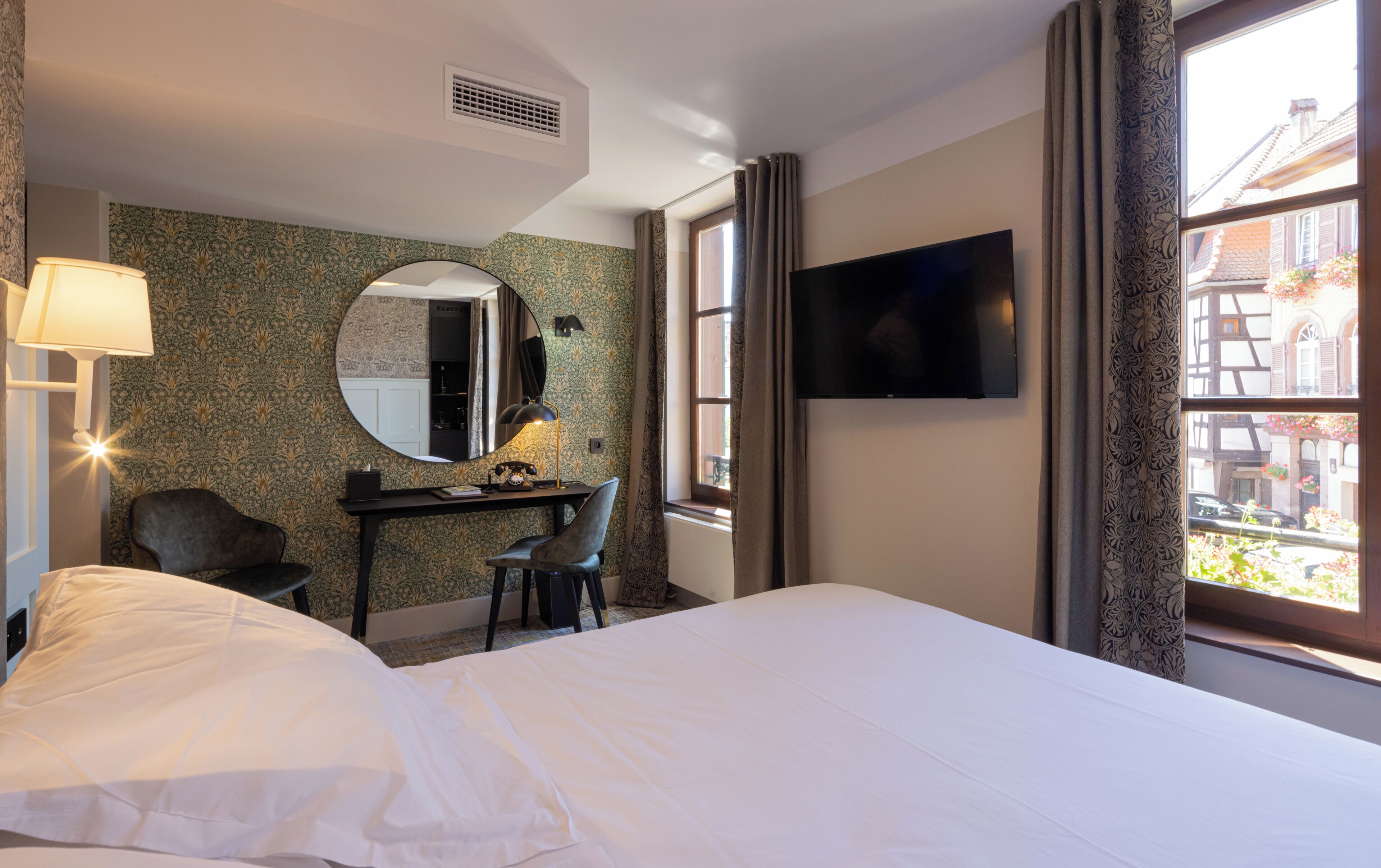 Ein Hotelzimmer mit Bett, Schreibtisch, Stuhl, Spiegel, Lampe, Fernseher und einem fensterbelaubten Fenster.