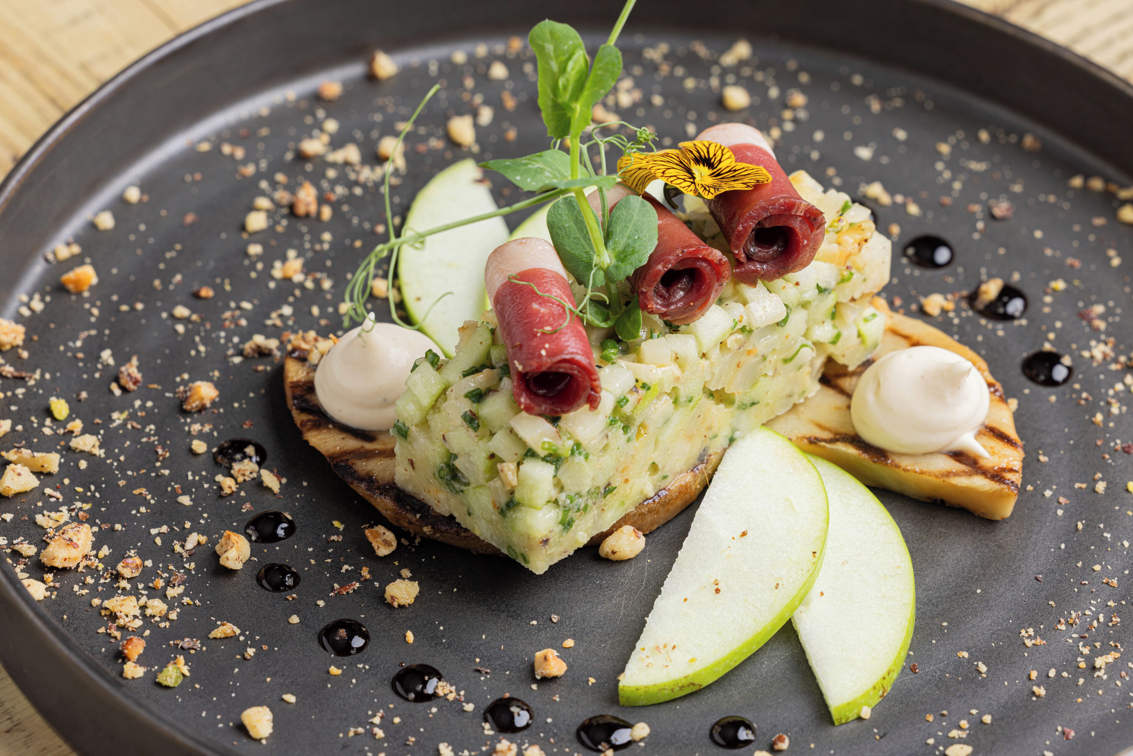 A dish with a slice of bread topped with a green mixture, a slice of apple, and a dollop of cream.