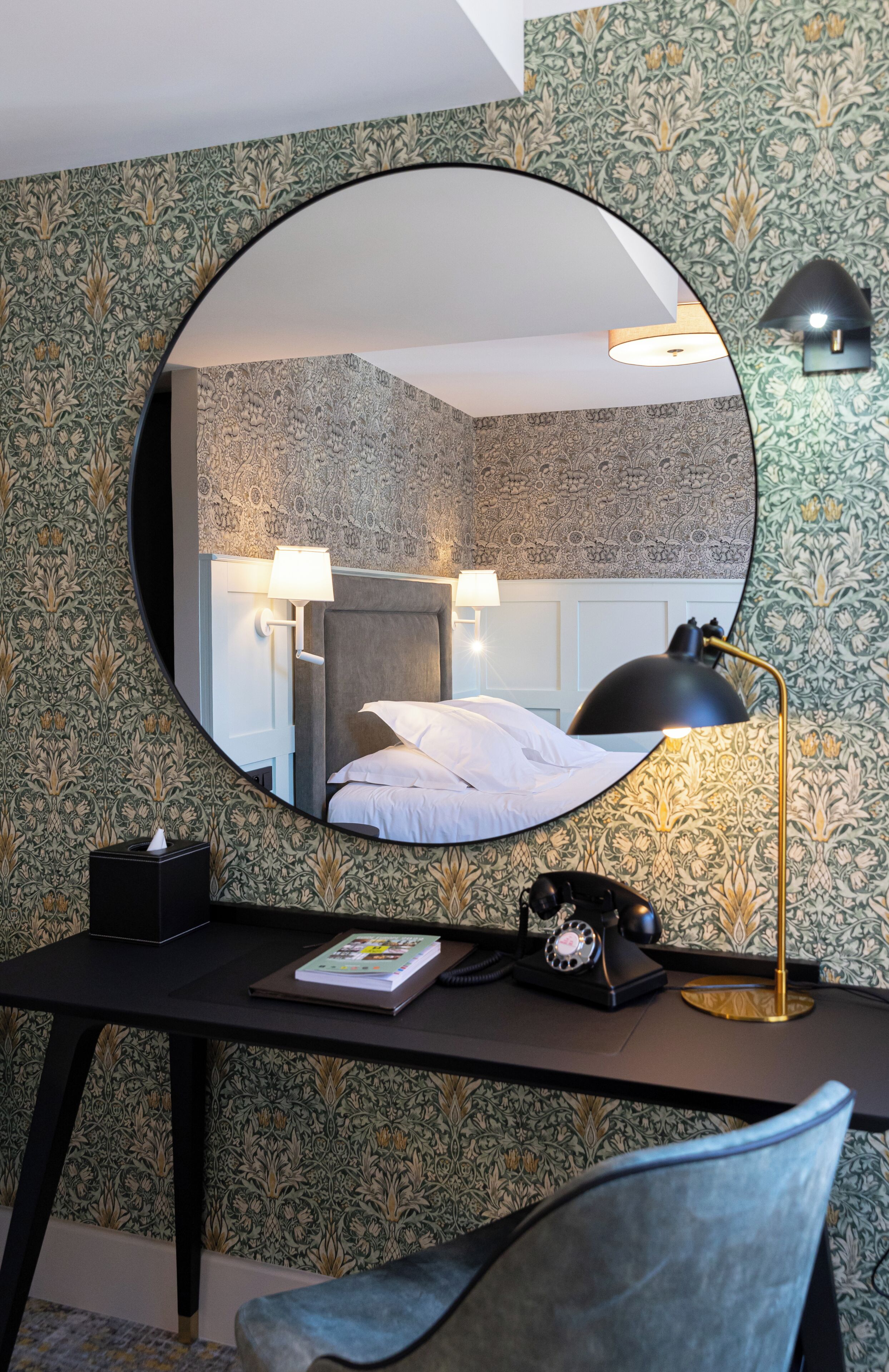 A hotel room with a round mirror reflecting a bed, a desk with a lamp, a telephone, and a book.
