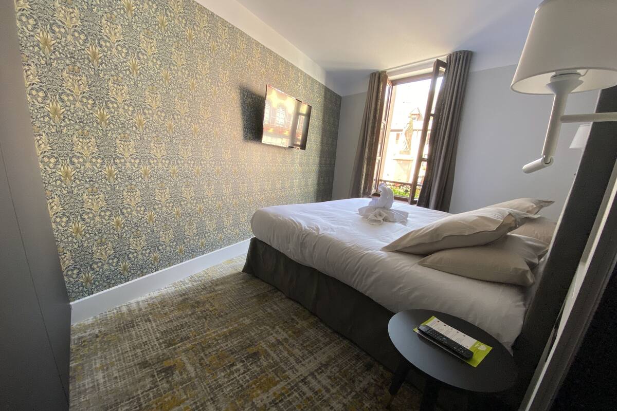 A hotel room with a bed, a small table, a lamp, and a window with curtains.