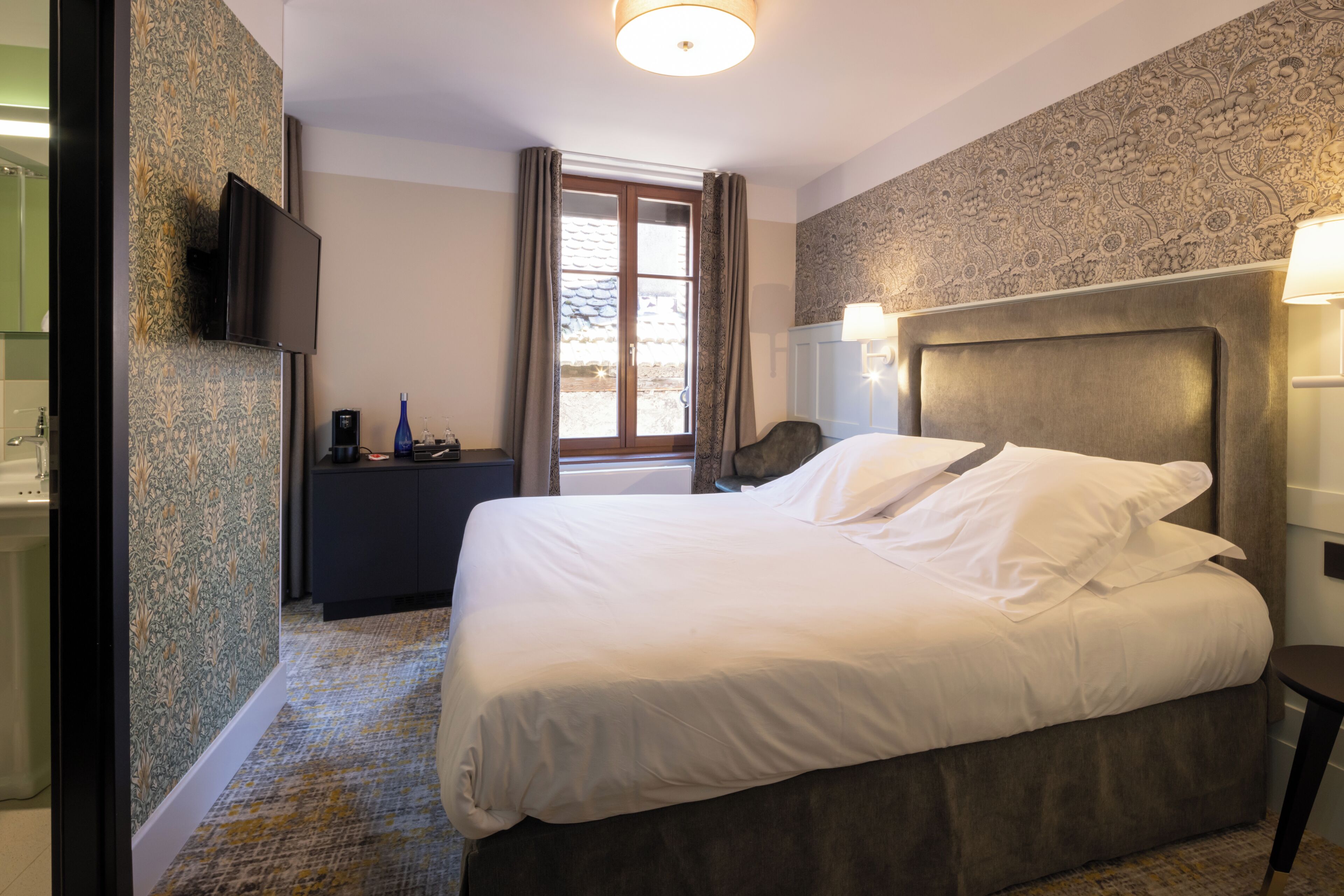 A hotel room with a bed, a television, a desk, a chair, a window with curtains, and a patterned wall.