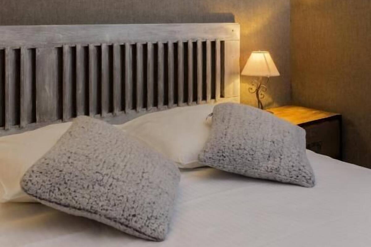 A hotel room with a modern design, featuring a large bed with a grey headboard, two grey pillows, a bedside table with a lamp, and a wall-mounted light fixture.