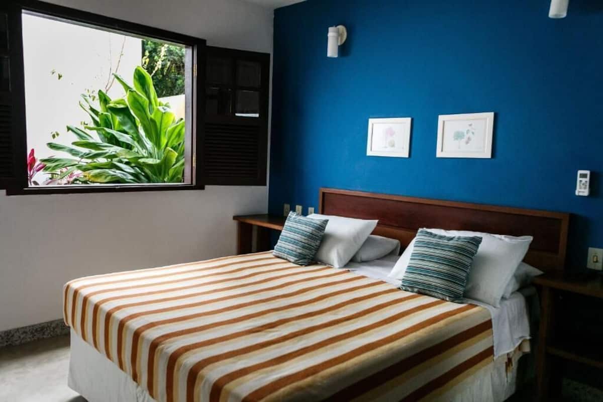A bed with striped bedding, a window with plants, and two framed pictures on a blue wall.