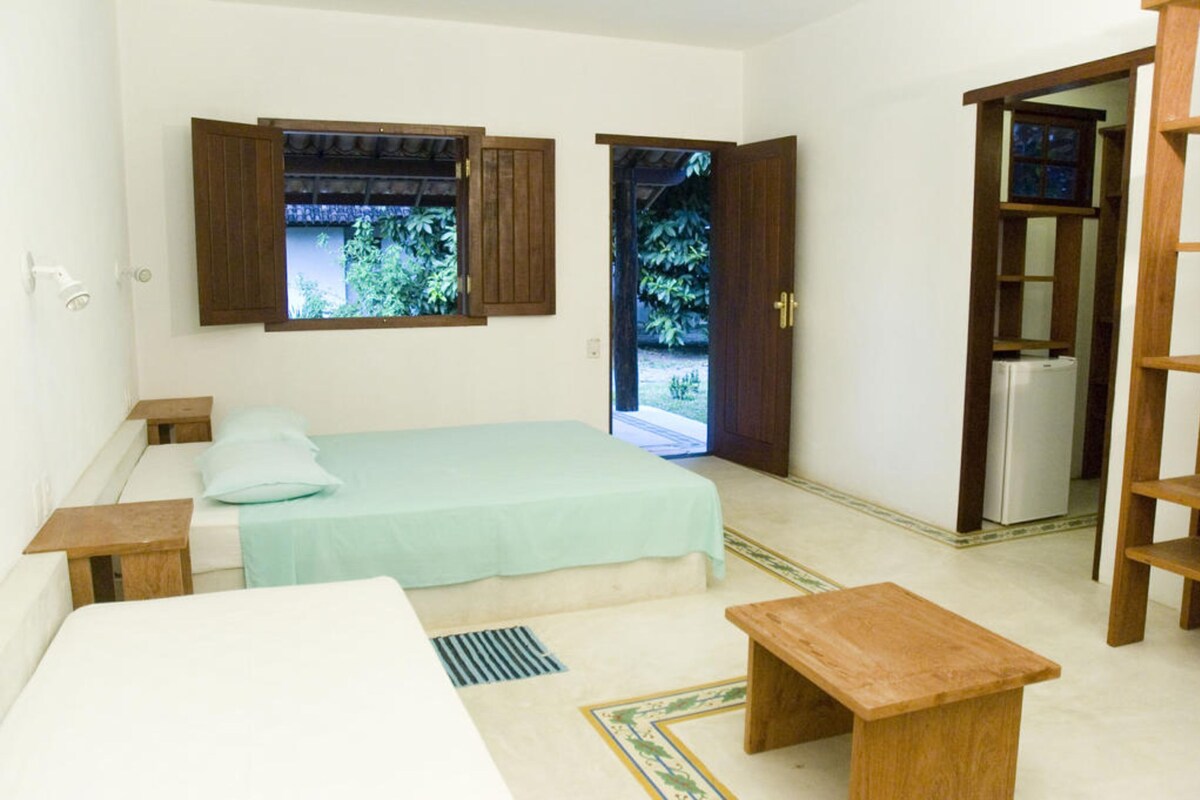 A room with a bed, a wooden bench, a small table, a window with shutters, and a door leading outside.