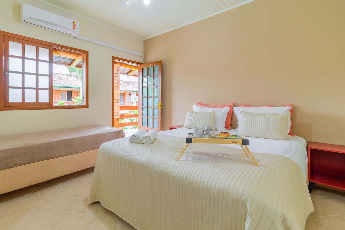 A hotel room with a bed, a bench, a window with a view, and an air conditioning unit.